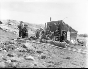 Image: Six men sitting by hut with coffee cups. Skins hanging